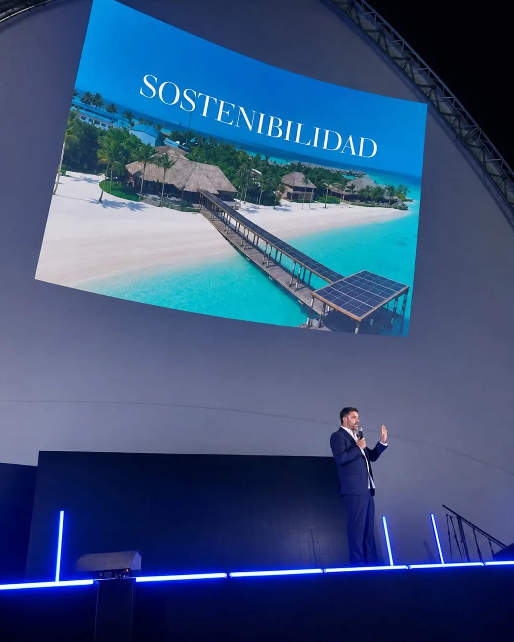 Héctor De Castro speaking on stage beneath a large screen showing a beach resort with the word Sostenibilidad
