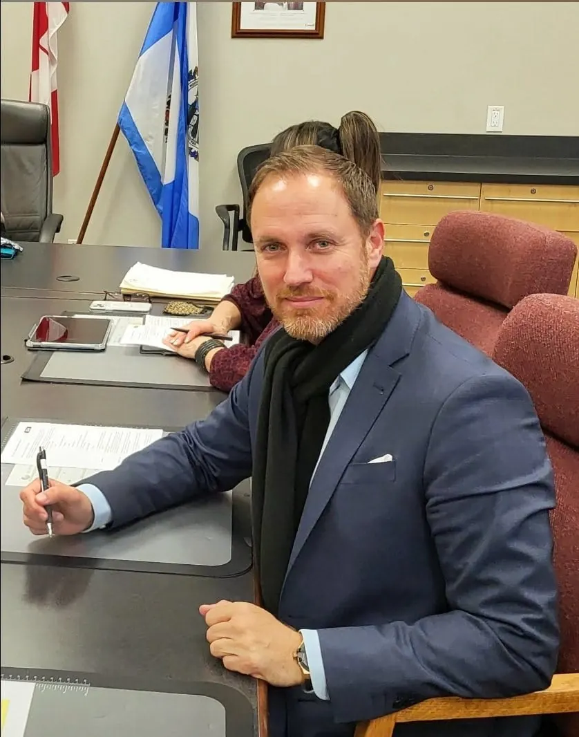 Héctor De Castro signing documents at the council table of Kelowna City Hall, Canada, with Canadian and Kelowna flags behind